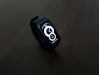 A digital smartwatch with a rectangular face displaying the time and date in a bright white interface. The watch is set against a dark, minimalistic background, highlighting its sleek and modern design.