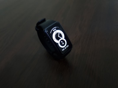 A digital smartwatch with a rectangular face displaying the time and date in a bright white interface. The watch is set against a dark, minimalistic background, highlighting its sleek and modern design.