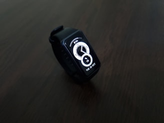 A digital smartwatch with a rectangular face displaying the time and date in a bright white interface. The watch is set against a dark, minimalistic background, highlighting its sleek and modern design.
