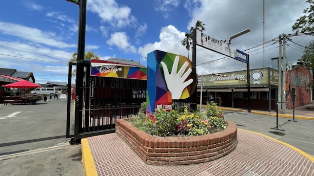 A colorful sign featuring a large hand icon is positioned in a circular brick planter filled with flowers. The surrounding area includes a paved street with an entrance sign overhead and a wide expanse of sky with scattered clouds. Nearby buildings have signage, and there are seating areas with red umbrellas.