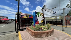 A colorful sign featuring a large hand icon is positioned in a circular brick planter filled with flowers. The surrounding area includes a paved street with an entrance sign overhead and a wide expanse of sky with scattered clouds. Nearby buildings have signage, and there are seating areas with red umbrellas.