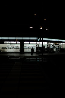 A dimly lit street scene at night featuring a FamilyMart convenience store. The store is illuminated, displaying its interior and signage. In front of the store, a silhouette of a person on a bicycle is visible against the light from the store. The foreground consists of a dark, empty street with pedestrian crossings barely visible.