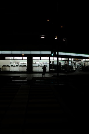 A dimly lit street scene at night featuring a FamilyMart convenience store. The store is illuminated, displaying its interior and signage. In front of the store, a silhouette of a person on a bicycle is visible against the light from the store. The foreground consists of a dark, empty street with pedestrian crossings barely visible.