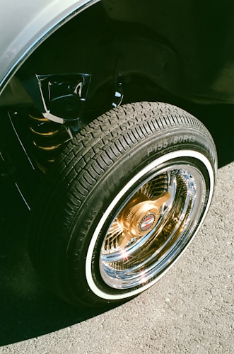 A close-up view of a car's wheel with sparkling chrome rims and a tire featuring distinctive white sidewalls. The sunlight reflects brightly off the metallic surface, creating a shiny appearance.