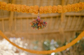 A decorative arrangement featuring a garland of vibrant orange marigold flowers draped against a blurred thatched background. In the center hangs a colorful tassel made from various fibers, creating a festive atmosphere.