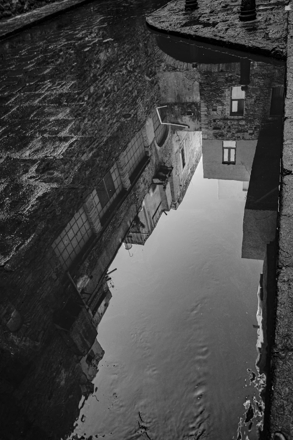 The image shows a reflection of a building and its surroundings captured in black and white, creating an interesting visual effect. The building's reflection is visible on the water below it, adding depth to the scene. The perspective of the photo is taken from above, looking down at the building and its reflection.