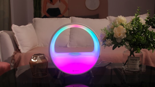 A modern, circular mood lamp emitting a gradient of colors, including blue, pink, and white, sits on a glass table. It is surrounded by a small glass candle holder and a vase filled with white and green flowers. In the background, there is a white couch with plush cushions, and a decorative art piece on the wall. The setting suggests a cozy and elegantly furnished living room.