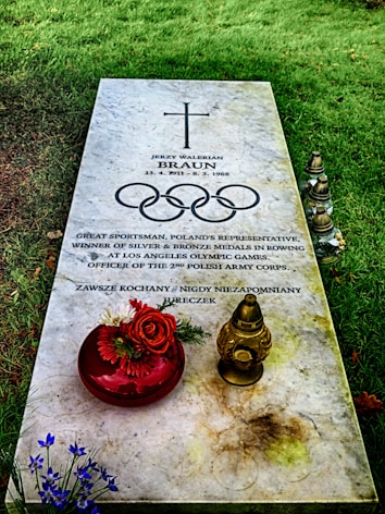 A gravestone set in a grassy area, inscribed with the name Jerzy Walerian Braun and dates 1911 to 1968. It celebrates his achievements as a sportsman, noting his medals in rowing at the Los Angeles Olympic Games. An arrangement of flowers in a red vase and a decorative candle holder rest on the stone.