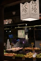 A market stall displaying an array of fresh fruits arranged in trays. Overhead, a sign with Chinese characters and 'SHUANGTA MARKET' printed on it is visible. The ambient lighting creates a warm and somewhat dim atmosphere, with other parts of the market vaguely visible in the background.