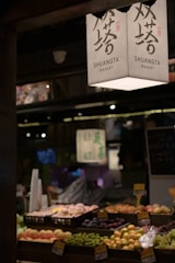 A market stall displaying an array of fresh fruits arranged in trays. Overhead, a sign with Chinese characters and 'SHUANGTA MARKET' printed on it is visible. The ambient lighting creates a warm and somewhat dim atmosphere, with other parts of the market vaguely visible in the background.