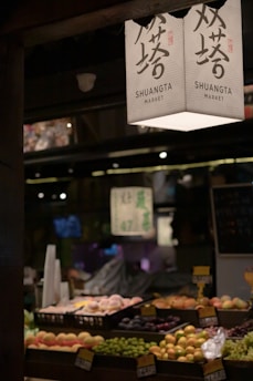A market stall displaying an array of fresh fruits arranged in trays. Overhead, a sign with Chinese characters and 'SHUANGTA MARKET' printed on it is visible. The ambient lighting creates a warm and somewhat dim atmosphere, with other parts of the market vaguely visible in the background.