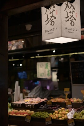 A market stall displaying an array of fresh fruits arranged in trays. Overhead, a sign with Chinese characters and 'SHUANGTA MARKET' printed on it is visible. The ambient lighting creates a warm and somewhat dim atmosphere, with other parts of the market vaguely visible in the background.
