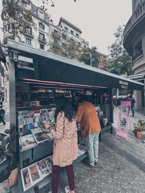 A newsstand situated on a city street, offering a variety of magazines and newspapers. Two people are browsing through the selection. The stand's awning is black with the words 'News & Coffee'. Surrounding the kiosk are tall buildings and trees, indicating an urban setting. The pavement is patterned, and there are potted plants nearby.