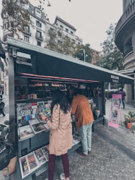 A newsstand situated on a city street, offering a variety of magazines and newspapers. Two people are browsing through the selection. The stand's awning is black with the words 'News & Coffee'. Surrounding the kiosk are tall buildings and trees, indicating an urban setting. The pavement is patterned, and there are potted plants nearby.