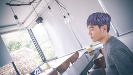 A person with purple hair is sitting at a long table in a modern, bright room with large windows and hanging light bulbs. The person appears to be in a relaxed state, with closed eyes. The room has a minimalistic decor, featuring blinds and some greenery visible through the windows.