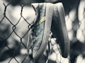 A pair of worn sneakers hangs by their laces on a chain-link fence, appearing slightly faded and weathered. The shoes feature decorative buttons in blue and yellow, adding a touch of color to their otherwise muted appearance. The background is blurred, focusing attention on the sneakers and the pattern of the fence.