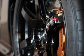 A close-up of a Nissan car wheel, featuring a black alloy rim with a prominently displayed red and white Nismo logo. The brake caliper is visible and painted in a metallic orange color, with the Nissan branding clearly printed in black.