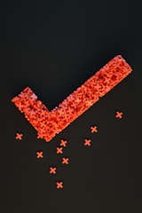 An arrangement of numerous small red crosses forms a large checkmark shape against a dark background, with several individual crosses scattered below.