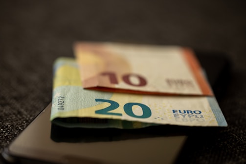 A close-up view of two Euro banknotes placed on top of a smartphone, with a 20 Euro note in the foreground and a 10 Euro note in the background. The background is out of focus, emphasizing the currency and the phone.