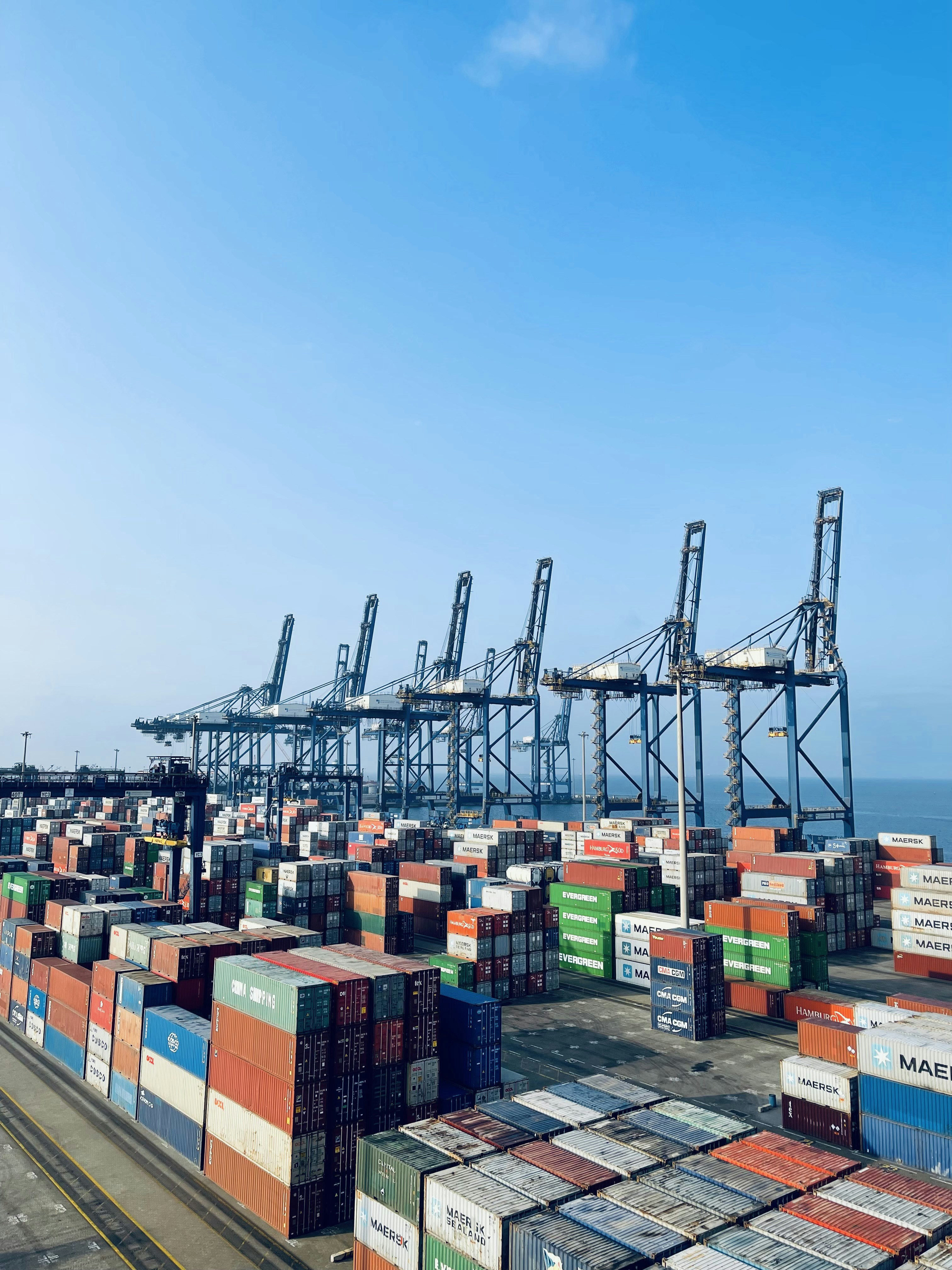 A busy shipping dock with containers being loaded onto a cargo ship under a clear sky.