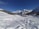 A snowy landscape with tracks leading towards a small village nestled at the base of a range of majestic, snow-capped mountains. Several people are skiing or walking in the snow, enjoying a clear, sunny day. Evergreen trees border the area, and the sky is a vibrant blue.