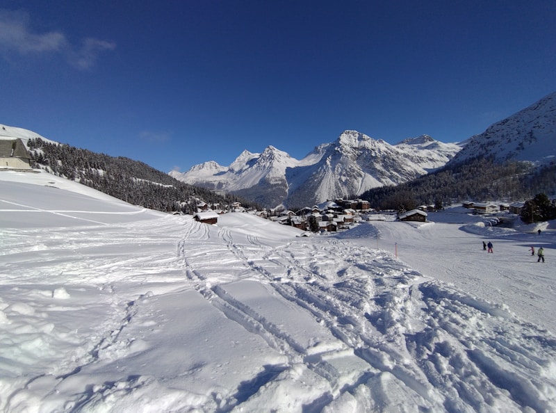 A snowy landscape with tracks leading towards a small village nestled at the base of a range of majestic, snow-capped mountains. Several people are skiing or walking in the snow, enjoying a clear, sunny day. Evergreen trees border the area, and the sky is a vibrant blue.