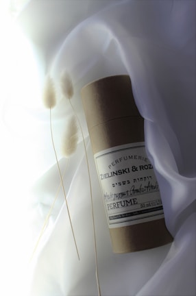 A cylindrical container labeled 'Zielinski & Rozen Perfume' rests on a soft, white fabric background. Delicate, dried pampas grass stems are placed beside it, adding an organic touch. The overall composition appears minimalist and elegant.