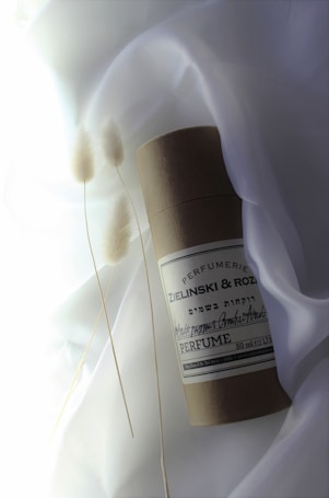 A cylindrical container labeled 'Zielinski & Rozen Perfume' rests on a soft, white fabric background. Delicate, dried pampas grass stems are placed beside it, adding an organic touch. The overall composition appears minimalist and elegant.