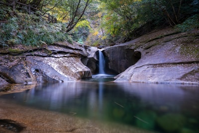 A serene landscape featuring a small waterfall cascading into a calm, clear pool surrounded by smooth, rocky formations. Lush green foliage and trees frame the scene, contributing to the peaceful ambiance.