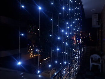 A balcony is decorated with strings of bright white LED lights, creating a sparkling curtain effect. The lights illuminate the surrounding area in a striking contrast with the night sky. Below, some buildings display additional colorful lights in the background, suggestive of a festive occasion. The balcony has a metal railing and a plastic chair is seated in one corner.