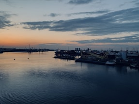 A calm harbor at dusk with moored cargo ships and cranes silhouetted against a navy blue sky.