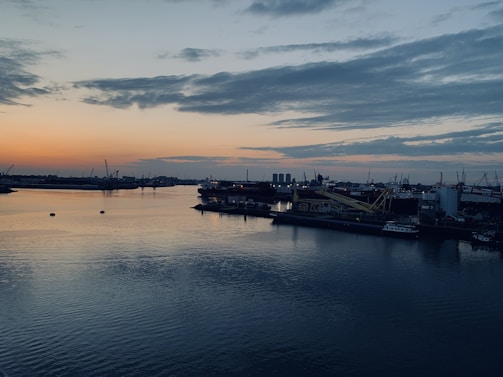 A calm harbor at dusk with moored cargo ships and cranes silhouetted against a navy blue sky.