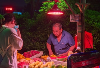 A street vendor dressed in a blue shirt stands behind a table laden with fresh fruits including bananas, oranges, and dragon fruit. Bright artificial lighting from a red overhead lamp casts a warm glow over the scene. A customer in a white shirt stands in front of the table, inspecting the items while holding a smartphone.