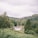 A serene rural landscape with lush green trees covering rolling hills. A quaint white cottage nestled among the greenery, surrounded by stone walls and small buildings. The sky is overcast, suggesting a calm, cloudy day.