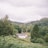 A serene rural landscape with lush green trees covering rolling hills. A quaint white cottage nestled among the greenery, surrounded by stone walls and small buildings. The sky is overcast, suggesting a calm, cloudy day.