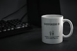 A white mug with the text 'HAPPINESS IS' printed in bold. Below the text, there is a humorous quote about having a younger brother who is taller, accompanied by a simple cartoon drawing of two characters. The mug is placed on a dark-colored surface next to a black computer keyboard.