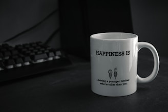 A white mug with the text 'HAPPINESS IS' printed in bold. Below the text, there is a humorous quote about having a younger brother who is taller, accompanied by a simple cartoon drawing of two characters. The mug is placed on a dark-colored surface next to a black computer keyboard.