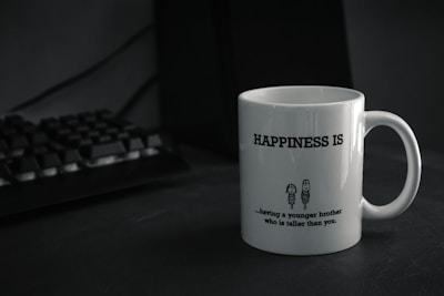A white mug with the text 'HAPPINESS IS' printed in bold. Below the text, there is a humorous quote about having a younger brother who is taller, accompanied by a simple cartoon drawing of two characters. The mug is placed on a dark-colored surface next to a black computer keyboard.