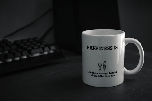 A white mug with the text 'HAPPINESS IS' printed in bold. Below the text, there is a humorous quote about having a younger brother who is taller, accompanied by a simple cartoon drawing of two characters. The mug is placed on a dark-colored surface next to a black computer keyboard.