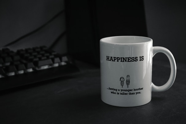 A white mug with the text 'HAPPINESS IS' printed in bold. Below the text, there is a humorous quote about having a younger brother who is taller, accompanied by a simple cartoon drawing of two characters. The mug is placed on a dark-colored surface next to a black computer keyboard.