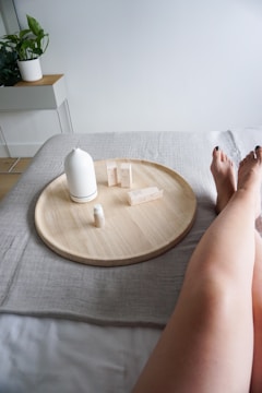 A pair of bare legs relaxed near a round wooden tray placed on a soft, beige fabric. On the tray are several small skincare or cosmetic items along with a white ceramic vase or bottle. In the background, there's a small green plant on a light-colored wooden table against a plain wall.