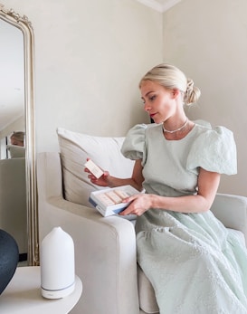 A woman wearing a light green dress sits on a cream-colored armchair, holding a box of skincare products. She is looking at one of the products in her hand. A large mirror with an ornate frame is visible in the background, and a white diffuser is placed on a small table beside the armchair.