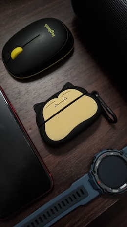 A wooden surface holds several items including a black and yellow Logitech wireless mouse, a smartphone with a red case, a unique Snorlax-themed protective case for wireless earbuds, and a blue digital wristwatch with black accents. The arrangement feels organized and personal.