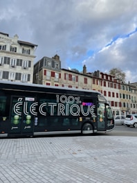 A modern electric bus is parked on a city street. The bus features large text on its side reading '100% électrique.' In the background, there are several traditional European buildings with colorful facades, some with red shutters. The sky above is cloudy with patches of blue visible.