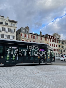 A modern electric bus is parked on a city street. The bus features large text on its side reading '100% électrique.' In the background, there are several traditional European buildings with colorful facades, some with red shutters. The sky above is cloudy with patches of blue visible.