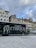 A modern electric bus is parked on a city street. The bus features large text on its side reading '100% électrique.' In the background, there are several traditional European buildings with colorful facades, some with red shutters. The sky above is cloudy with patches of blue visible.
