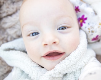 A close-up of a baby with light skin, wide eyes, and a slight smile. The baby is wearing a fluffy light-colored garment. A blanket with colorful floral patterns is visible in the background, adding a soft and cozy feel to the scene.