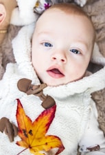 An infant is dressed in a fluffy, cream-colored garment with toggle buttons. A vibrant autumn leaf with shades of yellow, orange, and red rests on the baby's chest. The background features a soft brown texture, possibly a blanket or fur.