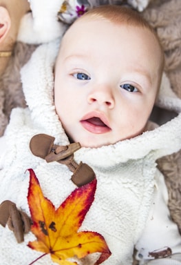 An infant is dressed in a fluffy, cream-colored garment with toggle buttons. A vibrant autumn leaf with shades of yellow, orange, and red rests on the baby's chest. The background features a soft brown texture, possibly a blanket or fur.