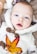 An infant is dressed in a fluffy, cream-colored garment with toggle buttons. A vibrant autumn leaf with shades of yellow, orange, and red rests on the baby's chest. The background features a soft brown texture, possibly a blanket or fur.